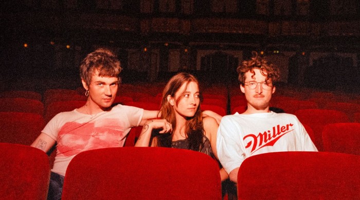 Three people sit in red theater seats, with the two on the sides leaning toward the person in the middle.